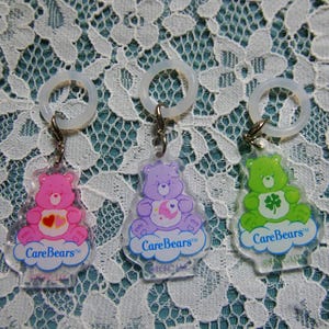Könnte beinhalten: Drei Care Bears Schlüsselanhänger. Ein rosa Bär mit einem Herz, ein lila Bär mit Mond und Sternen und ein grüner Bär mit einem vierblättrigen Kleeblatt. Jeder Schlüsselanhänger hat ein klares Kunststoff-Bär-Design, einen Metallverschluss und einen transparenten Kunststoffring.