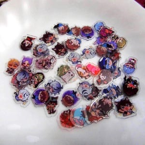 Könnte beinhalten: Eine Sammlung kleiner, klarer Acryl-Charms mit farbenfrohen Illustrationen von Charakteren im Anime-Stil. Die Charms haben verschiedene Designs, darunter Figuren mit unterschiedlichen Haarfarben und Outfits. Sie sind auf einer weißen Oberfläche verstreut.