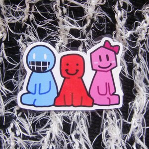 Puede incluir: Una pegatina con tres personajes de dibujos animados en azul, rojo y rosa. El personaje azul tiene una amplia sonrisa, el rojo sonr&iacute;e y el rosa lleva un lazo. La pegatina est&aacute; sobre un fondo texturizado en blanco y negro.