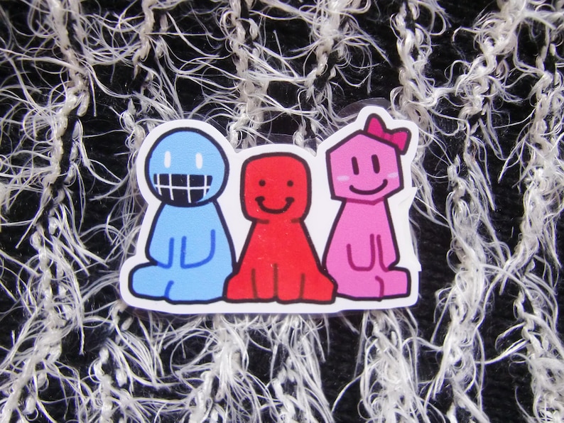 Puede incluir: Una pegatina con tres personajes de dibujos animados en azul, rojo y rosa. El personaje azul tiene una amplia sonrisa, el rojo sonr&iacute;e y el rosa lleva un lazo. La pegatina est&aacute; sobre un fondo texturizado en blanco y negro.