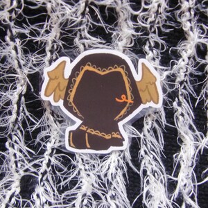 Puede incluir: Una pegatina con una figura marr&oacute;n con capucha con alas color canela y un detalle naranja. La pegatina est&aacute; sobre un fondo texturizado en blanco y negro. La figura tiene un borde decorativo.