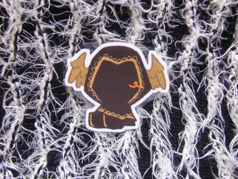 Puede incluir: Una pegatina con una figura marr&oacute;n con capucha con alas color canela y un detalle naranja. La pegatina est&aacute; sobre un fondo texturizado en blanco y negro. La figura tiene un borde decorativo.