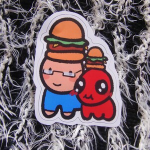 Puede incluir: Una pegatina con dos personajes de dibujos animados con hamburguesas en la cabeza. Un personaje es una persona con gafas y pantalones cortos azules, y el otro es una criatura roja. La pegatina tiene un borde blanco.