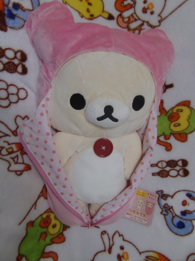 Puede incluir: Un oso de peluche blanco que lleva un saco de dormir con capucha rosa con lunares blancos. El oso tiene ojos negros y una nariz de bot&oacute;n. El saco de dormir tiene una cremallera y una etiqueta que dice "Rilakkuma".