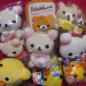 Op de afbeelding: Een verzameling pluche speelgoed met beren- en eendenfiguren. De beren zijn voornamelijk wit met roze accenten, en de eend is geel. Sommige speelgoed is verpakt in doorzichtig plastic.