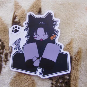 Puede incluir: Una pegatina troquelada con un personaje de dibujos animados con orejas de gato, pelo negro y un atuendo morado. El personaje tiene una expresi&oacute;n juguetona con la lengua fuera. Tambi&eacute;n se incluye una peque&ntilde;a pegatina con una huella de pata.