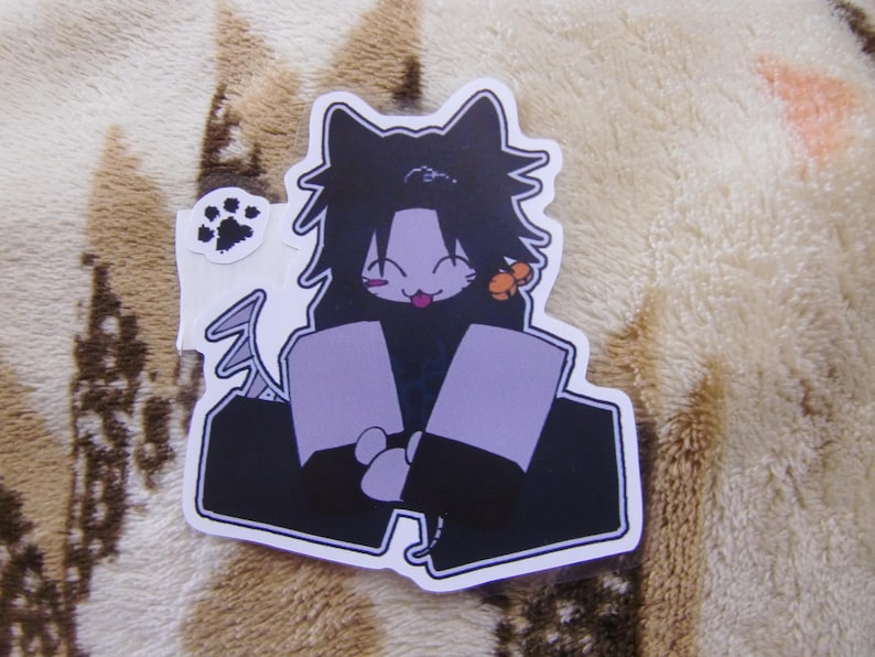 Puede incluir: Una pegatina troquelada con un personaje de dibujos animados con orejas de gato, pelo negro y un atuendo morado. El personaje tiene una expresi&oacute;n juguetona con la lengua fuera. Tambi&eacute;n se incluye una peque&ntilde;a pegatina con una huella de pata.