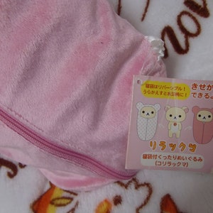 Puede incluir: Saco de dormir de felpa rosa con cremallera y una etiqueta que dice "El saco de dormir es reversible! &iexcl;Dale la vuelta y es de lunares!" y "&iexcl;C&aacute;mbiate de ropa!" en japon&eacute;s. La etiqueta tambi&eacute;n muestra tres ilustraciones de un oso con diferentes sacos de dormir. La etiqueta dice "Rilakkuma" y "Peluche saco de dormir" en japon&eacute;s.