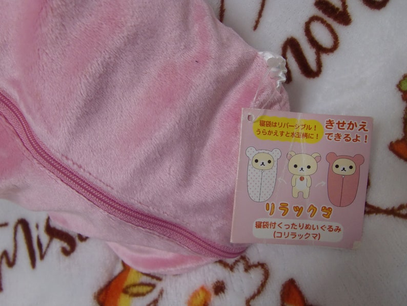 Puede incluir: Saco de dormir de felpa rosa con cremallera y una etiqueta que dice "El saco de dormir es reversible! &iexcl;Dale la vuelta y es de lunares!" y "&iexcl;C&aacute;mbiate de ropa!" en japon&eacute;s. La etiqueta tambi&eacute;n muestra tres ilustraciones de un oso con diferentes sacos de dormir. La etiqueta dice "Rilakkuma" y "Peluche saco de dormir" en japon&eacute;s.