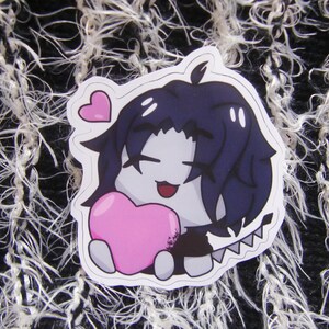 Puede incluir: Una pegatina con un personaje de estilo anime con cabello azul oscuro, sosteniendo un coraz&oacute;n rosa. El personaje tiene una expresi&oacute;n sonriente y est&aacute; sobre un borde blanco. Un peque&ntilde;o coraz&oacute;n rosa flota sobre la cabeza del personaje.