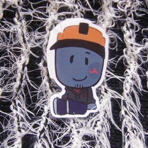 Puede incluir: Una pegatina con un personaje de dibujos animados con piel azul, que lleva un casco naranja y una sudadera con capucha gris. El personaje tiene una peque&ntilde;a marca roja en la mejilla y est&aacute; sentado. El fondo es una tela texturizada en blanco y negro.