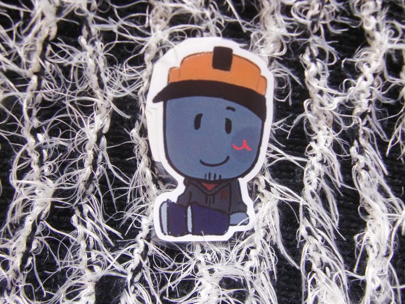 Puede incluir: Una pegatina con un personaje de dibujos animados con piel azul, que lleva un casco naranja y una sudadera con capucha gris. El personaje tiene una peque&ntilde;a marca roja en la mejilla y est&aacute; sentado. El fondo es una tela texturizada en blanco y negro.