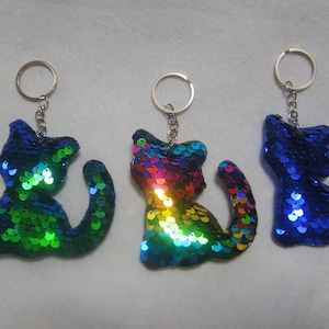 Op de afbeelding: Drie pailletten kat sleutelhangers in groen, regenboog en blauw. Elke kat zit met een gebogen staart en is bevestigd aan een zilveren sleutelhanger. De pailletten creëren een glinsterend effect.