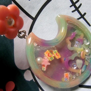 May include: A crescent moon-shaped resin pendant featuring a pastel rainbow gradient border. The center is filled with clear liquid, small white pearls, pink star charms, heart-shaped frames, and colorful fruit-themed polymer clay slices.