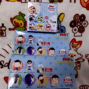 May include: Three boxes of Japanese anime character clip badges. The boxes are light blue with cartoon illustrations of the characters. The badges are round and feature various character designs. The boxes are stacked on a patterned blanket.