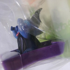 May include: A blue figurine of a cartoon character with a black cape and hat, sitting in a purple coffin-shaped boat. The figurine has a red tongue and is packaged in clear plastic.