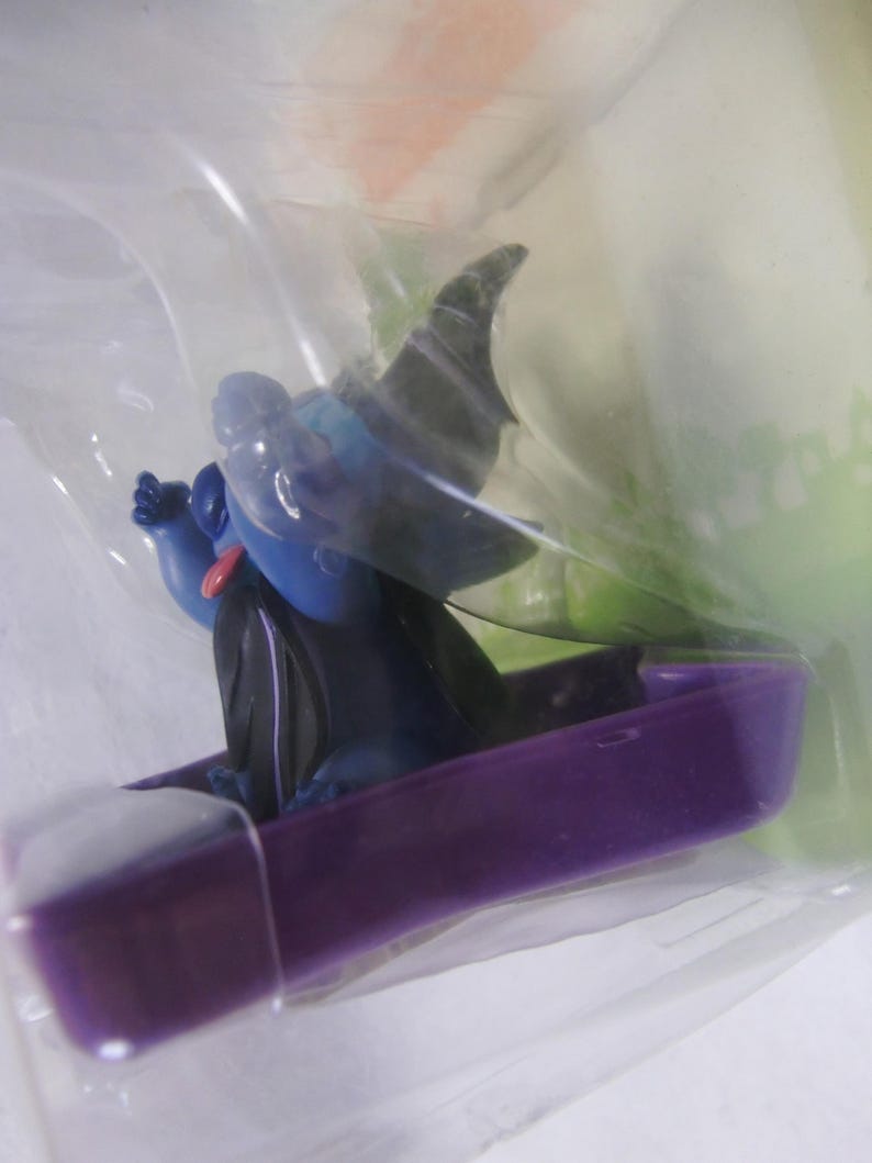 May include: A blue figurine of a cartoon character with a black cape and hat, sitting in a purple coffin-shaped boat. The figurine has a red tongue and is packaged in clear plastic.