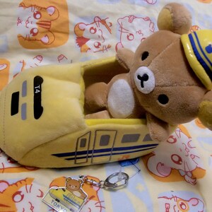 Pu&ograve; includere: Un giocattolo di peluche giallo a forma di treno proiettile giapponese con un orso marrone che indossa un cappello blu e giallo seduto all'interno. Il treno ha il numero T4 sul lato e le parole "TOKYO" e "JAPAN" in basso.