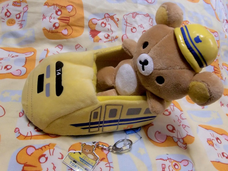 Pu&ograve; includere: Un giocattolo di peluche giallo a forma di treno proiettile giapponese con un orso marrone che indossa un cappello blu e giallo seduto all'interno. Il treno ha il numero T4 sul lato e le parole "TOKYO" e "JAPAN" in basso.