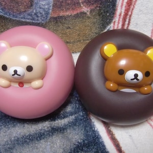 May include: Two donut-shaped plush toys with bear faces. The pink donut has a light brown bear with a red heart on its chest. The brown donut has a brown bear with yellow ears.