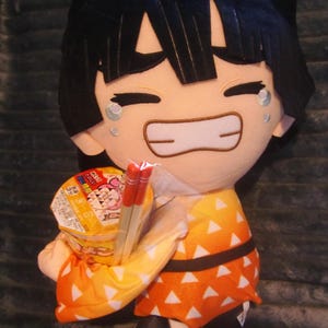 Pode incluir: Boneca de pelúcia de um personagem com cabelo preto e uma roupa estampada em amarelo e laranja. A boneca está segurando um copo de macarrão instantâneo e um par de pauzinhos vermelhos. A boneca tem uma expressão feliz com lágrimas.