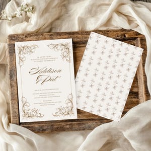 Op de afbeelding: Een witte huwelijksuitnodiging met een gouden, sierlijke rand en de namen Addison & Pat gedrukt in bruine cursieve letters. De uitnodiging ligt op een houten oppervlak naast een witte kaart met een patroon.