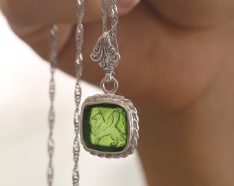 May include: A silver chain necklace with a green glass pendant featuring a detailed design. The pendant is square shaped with a silver border.