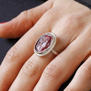 Può includere: Un anello d'argento con un cammeo rosso che presenta un profilo intagliato di una testa umana. L'anello è su un dito con smalto rosso.