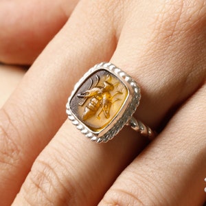 May include: A silver ring with a square-shaped amber stone featuring a carved bee design.