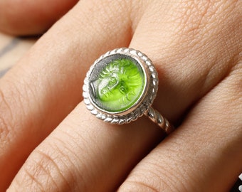 May include: A silver ring with a green glass cameo featuring a face. The ring has a twisted band and a beaded edge.