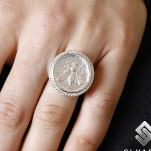 May include: A silver ring featuring a detailed bee design. The ring is worn on a finger.