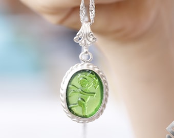 Puede incluir: Un collar de cadena de plata con un colgante de vidrio verde que presenta una talla detallada de una figura alada. El colgante tiene forma ovalada y tiene un borde plateado.