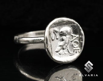 May include: A silver ring with a round, textured top featuring a detailed design of a winged figure. The ring is set against a black background.