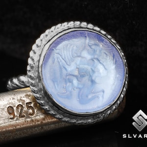 Könnte beinhalten: Ein silberner Ring mit einem blauen Glas-Kamee, der eine detaillierte Schnitzerei einer mythischen Kreatur zeigt. Der Ring ist mit "925" gekennzeichnet, was bedeutet, dass er aus Sterlingsilber besteht.