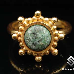 May include: A gold ring featuring a green gemstone set in a circular bezel. The ring is adorned with a series of small gold beads around the bezel.