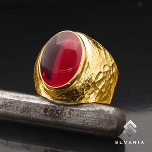 May include: A gold ring with a large, oval-shaped, red gemstone. The ring has a textured, hammered finish.