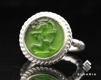 May include: A silver ring with a green glass cameo featuring a carved design. The ring has a rope-like band.