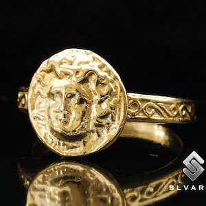 Puede incluir: Un anillo de oro con un diseño detallado de la cara de una mujer en la parte delantera. La banda del anillo está decorada con un patrón de espiral.