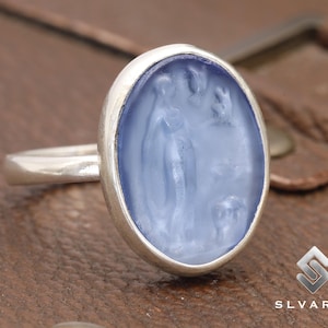 May include: A silver ring with a blue oval-shaped stone featuring a carved design of a woman and a bird.