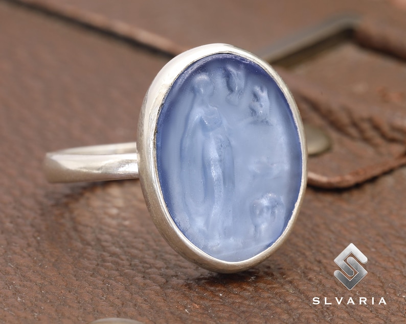May include: A silver ring with a blue oval-shaped stone featuring a carved design of a woman and a bird.