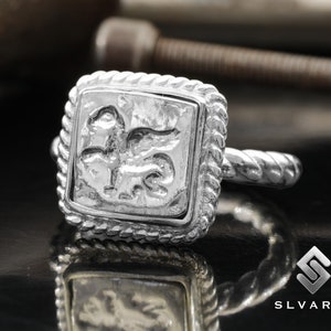 May include: A silver ring with a square design featuring a detailed embossed image of a mythical creature. The ring has a twisted band.