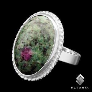 May include: A silver ring with an oval gemstone in shades of green, black, and pink. The stone is framed by a twisted rope design. The ring has a plain, polished band.