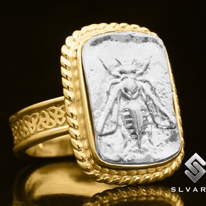 May include: A gold ring with a Celtic knot design on the band and a square silver inset featuring a detailed embossed image of a bee.