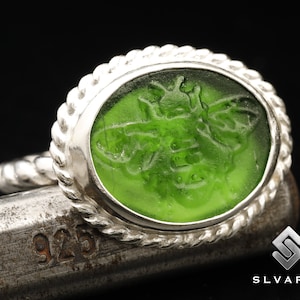 May include: A silver ring with a green glass oval inset featuring a detailed bee design. The ring has a rope-like band.