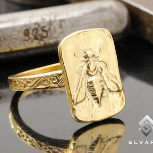 Può includere: Un anello d'oro con un designo dettagliato di un'ape sulla faccia dell'anello. La fascia dell'anello è decorata con un motivo a spirale.