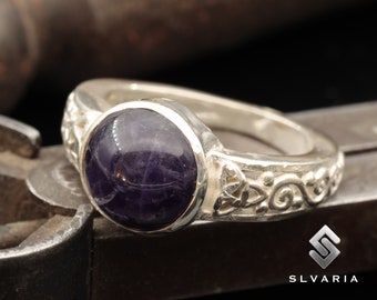 Peut inclure: Une bague en argent avec une pierre précieuse violette foncée sertie dans un sertissage à bélière. La bande présente un motif de nœud celtique et des volutes complexes.