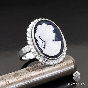 Peut inclure: Bague en argent avec un camée ovale représentant un profil blanc sur fond noir. La bague a un bord tressé et une bande polie. La bague est posée sur une surface métallique. La marque "SLVARIA" est visible.