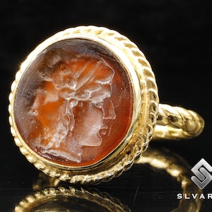 May include: A gold ring with a carved carnelian stone featuring a detailed profile of a woman's face. The ring has a rope-like design around the stone.