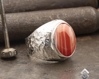 May include: A silver ring with a hammered texture and a red and white agate gemstone. The ring is set in a bezel setting.