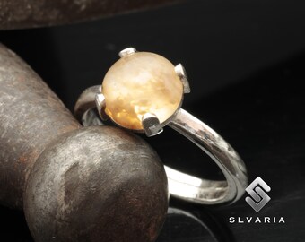 May include: A silver ring with a round, light brown gemstone set in a four-prong setting. The ring is on a dark, textured surface.